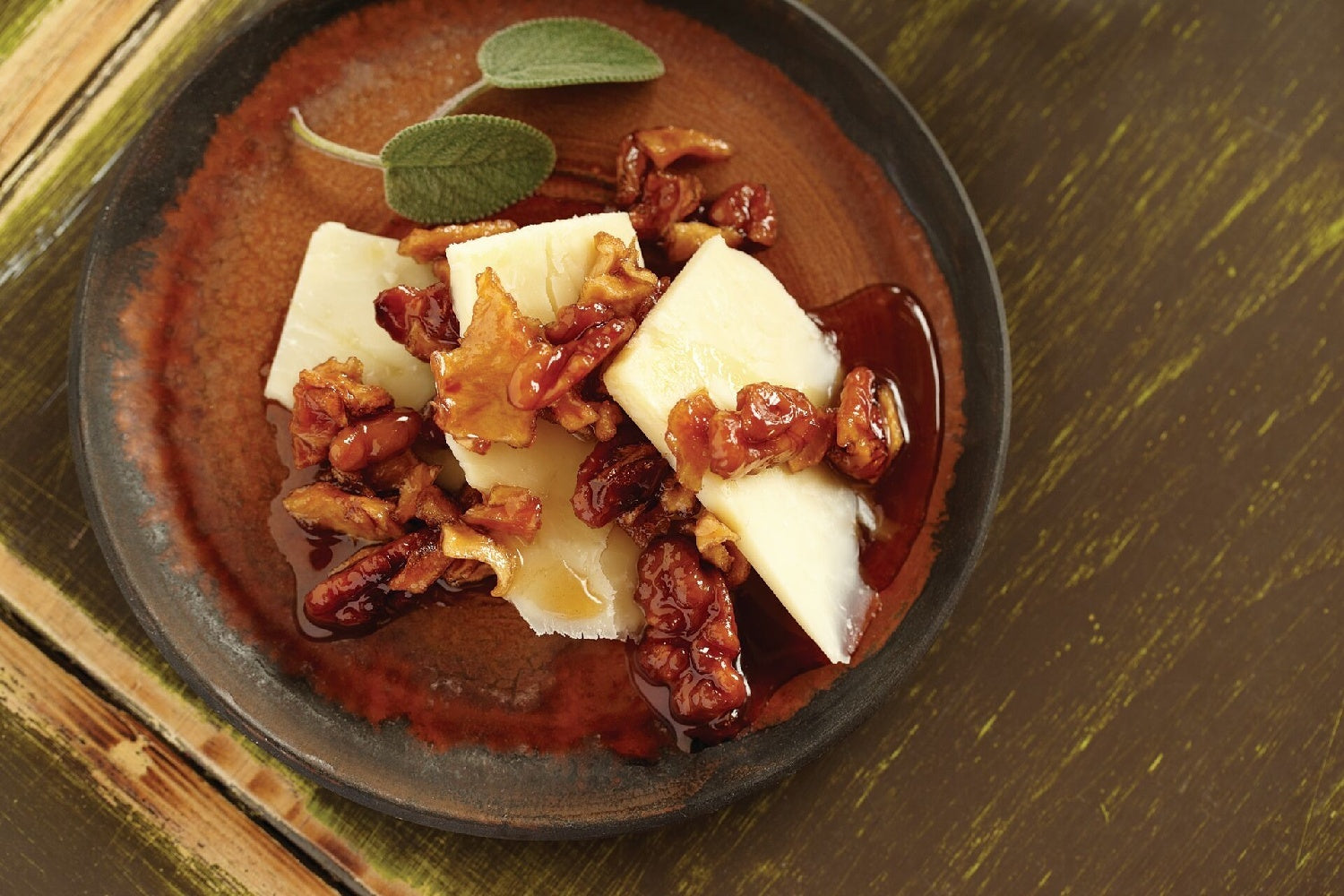 Cheese slices topped with glazed nuts and honey, garnished with fresh sage leaves on a rustic plate.